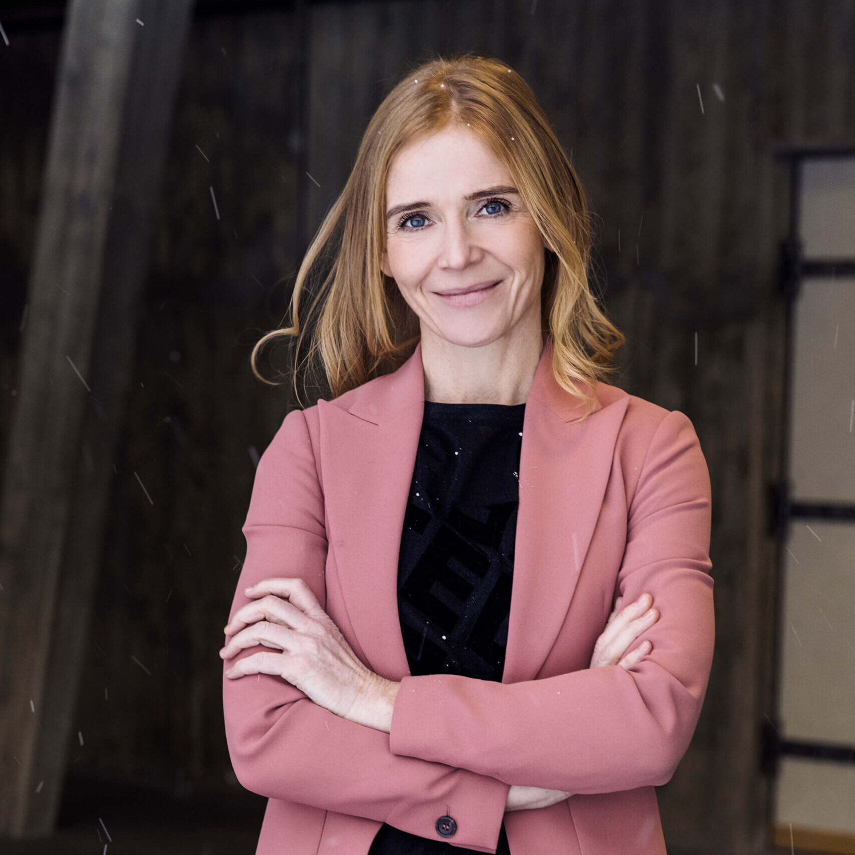 Porträtt på kvinna i jordgubbsblont hår, svart topp och rosa kavaj som står med armarna i kors och ler mot kameran