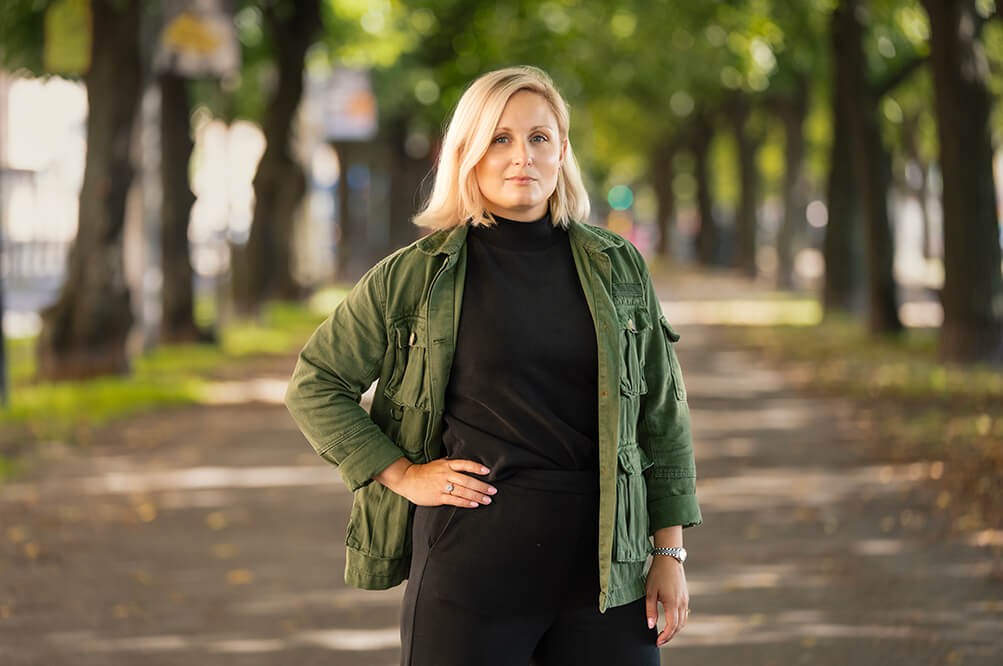 Porträtt på kvinna med blont axellångt hår, svarta kläder och grön jacka som står i utomhus